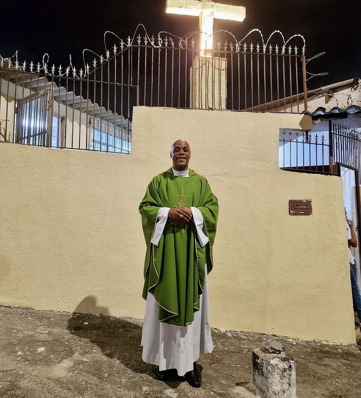 Padre diante do muro com a cruz iluminada, no espaço definitivo da Comunidade Nossa Senhora da Penha
